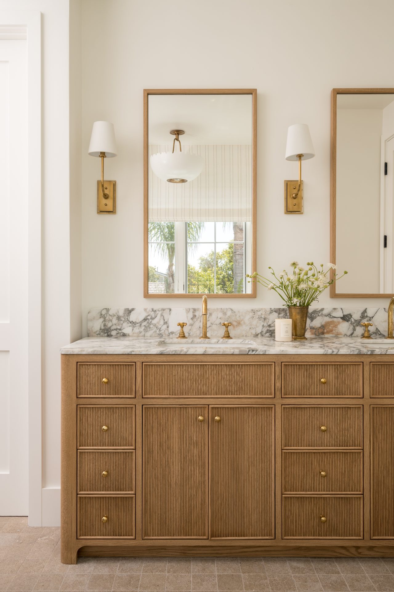 Bathroom vanity with natural wood cabinetry, marble counter, brass fixtures, and wall sconces
