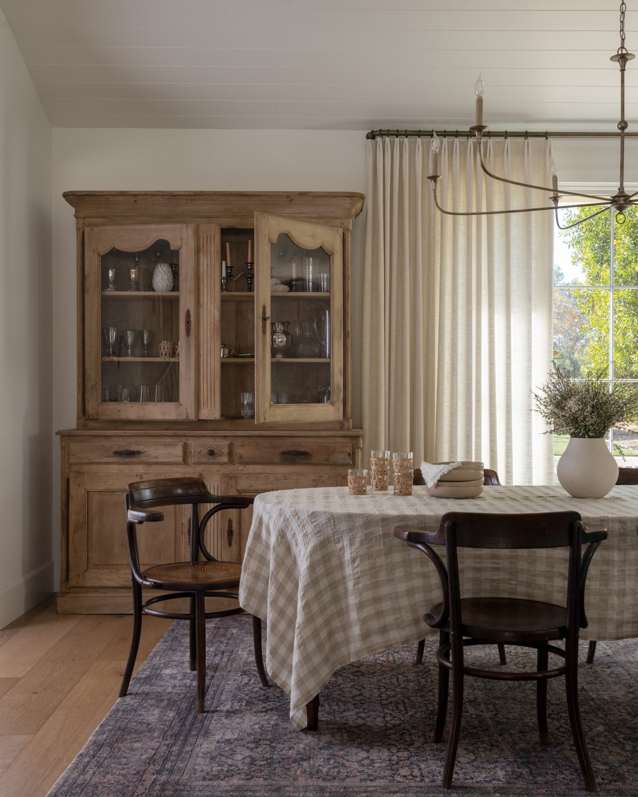Dining room with antique wood hutch, soft drapery, patterned rug, and layered table styling